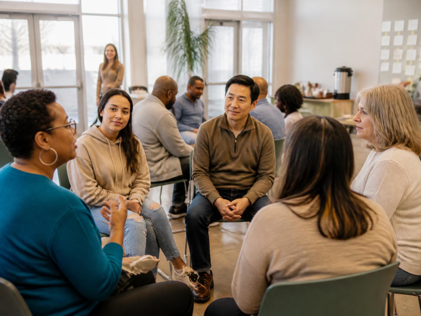 A diverse group of adults standing in small clusters, talking and listening to one another in a bright, daylit community hall